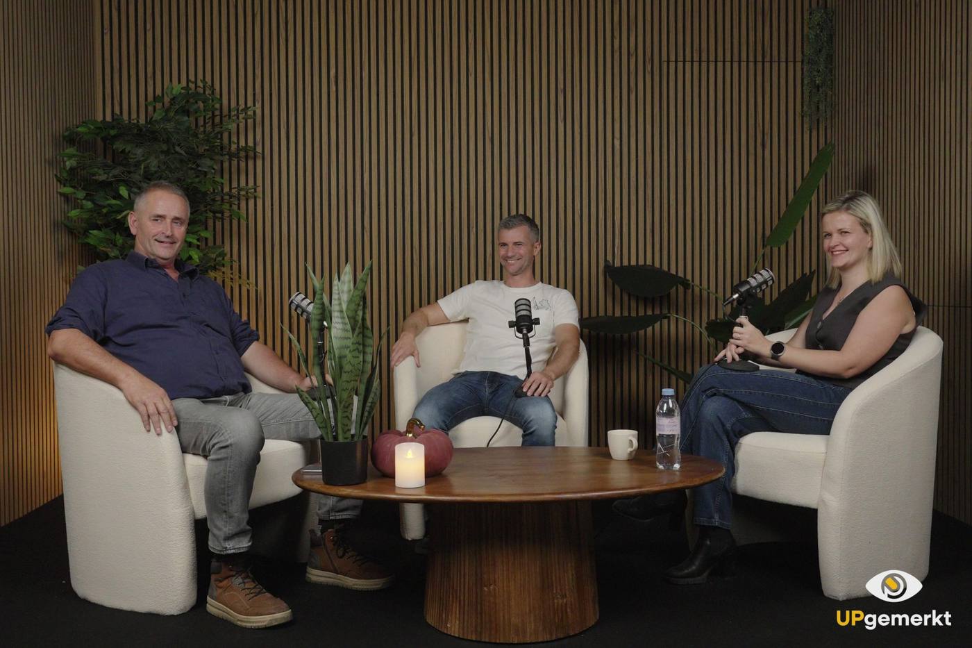 Drie mensen in gesprek in de studio: lichte zetels rond een lage houten tafel, microfoons op statief en een houten lattenwand met planten